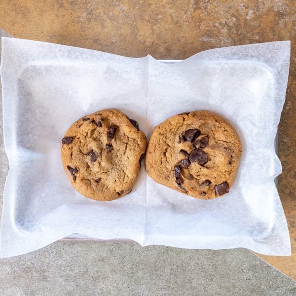 Two chocolate chip cookies on white parchment paper in a metal tray on a tiled surface.
