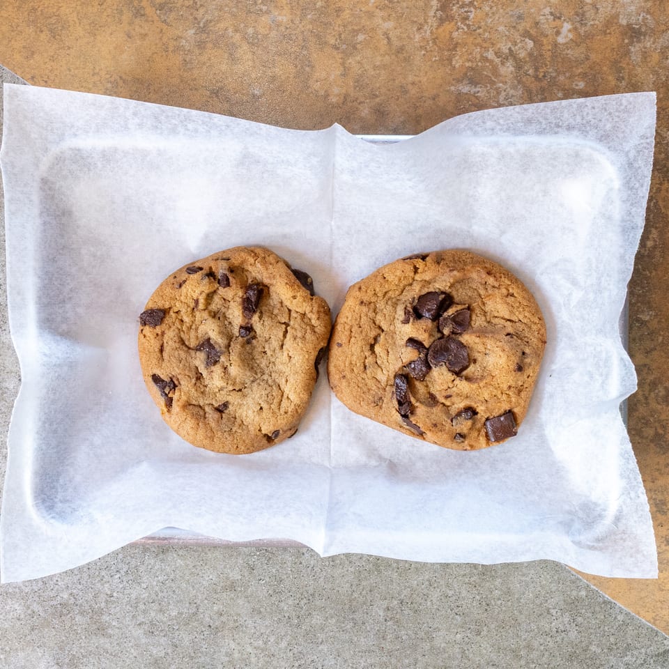 Two chocolate chip cookies on white parchment paper in a metal tray on a tiled surface.