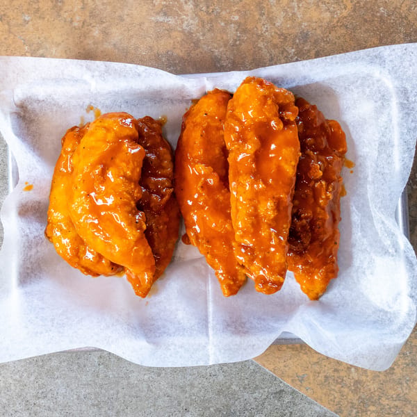 Four crispy chicken tenders coated in a vibrant orange buffalo sauce, served on white parchment paper.