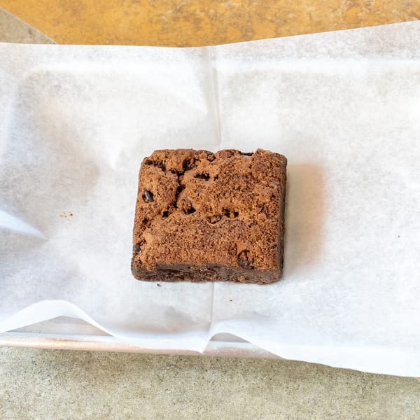 A single square chocolate brownie with a crumbly top sits on white parchment paper.