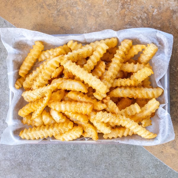 A metal tray filled with golden, crinkle-cut french fries seasoned with pepper, on a two-toned surface.